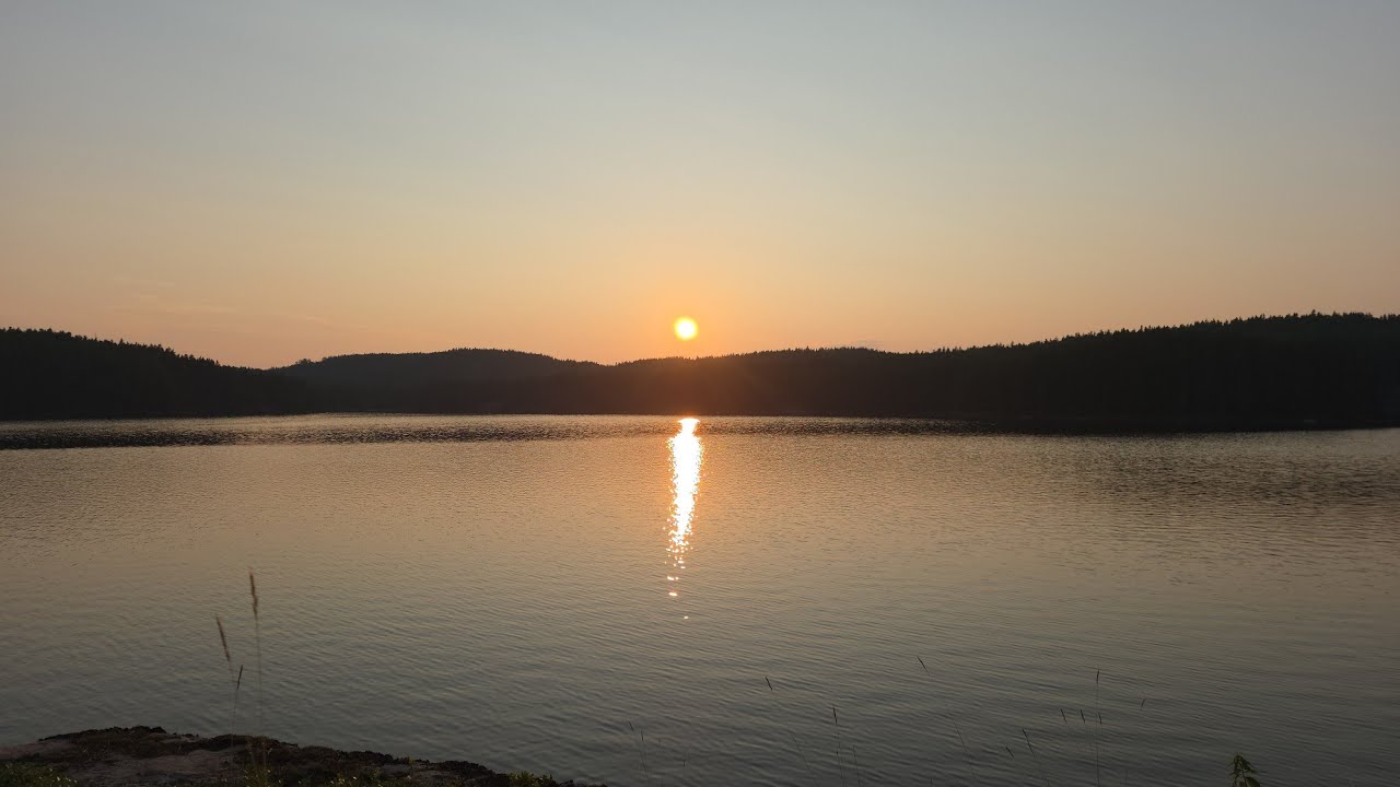 The sun setting over a calm lake ringed by dark forested hills