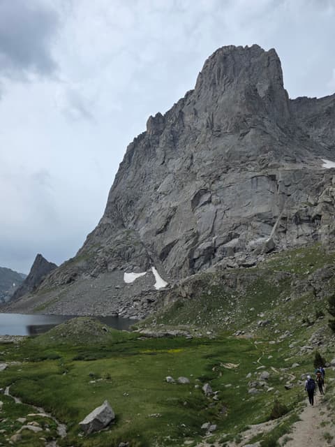 A granite spire above an alpine meadow and lake, with hikers on the trail