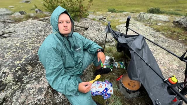 Finn in a teal rain suit cooking at a rocky campsite