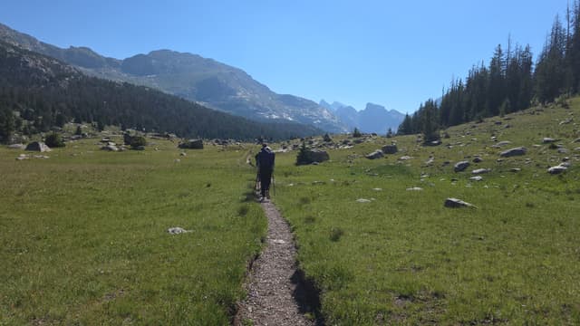 A hiker on a trail through a green meadow toward granite mountains