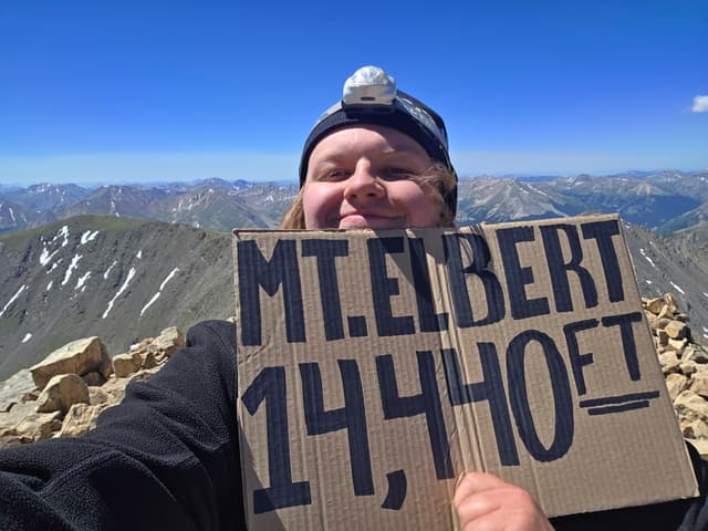 Finn holding a cardboard sign reading Mt. Elbert 14,440 ft on the summit
